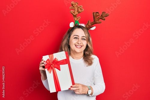 Middle age caucasian woman wearing cute christmas reindeer horns holding gift looking positive and happy standing and smiling with a confident smile showing teeth