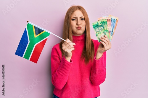 Young irish woman holding south africa flag and rand banknotes puffing cheeks with funny face. mouth inflated with air, catching air.