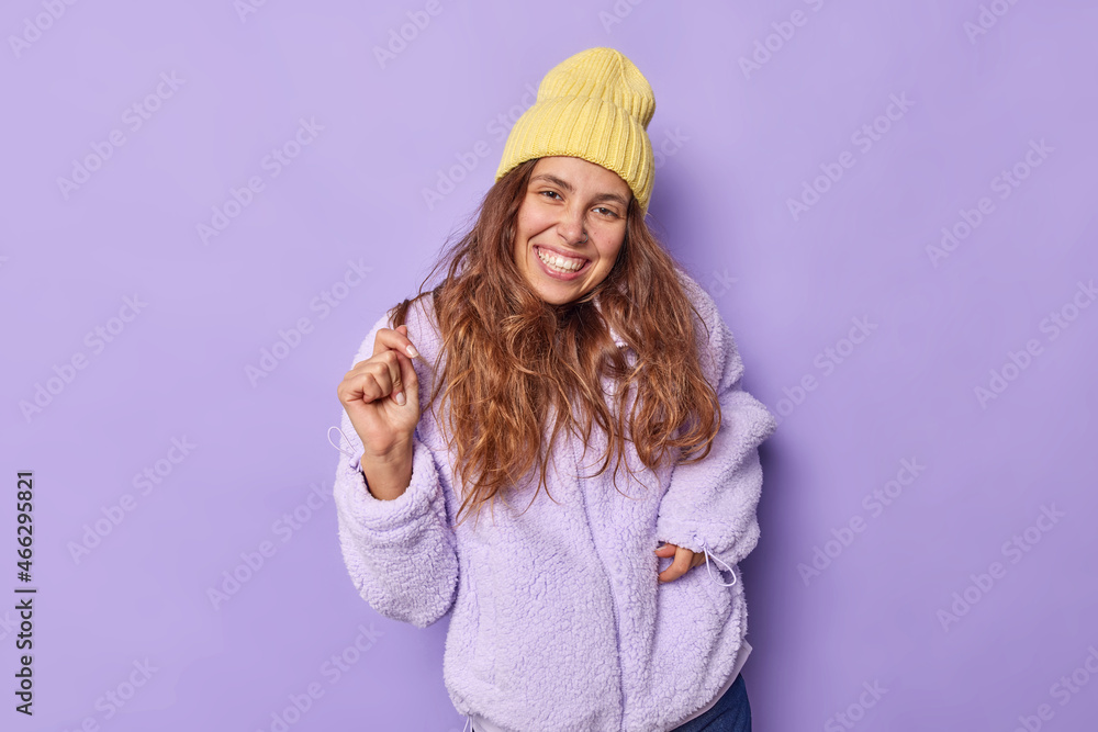 Photo of pretty brunette female model smiles broadly shows white teeth moves carefree dressed in fur coat hat on head enjoys life going to have walk during winter day isolated over purple wall Photo of pretty brunette female model smiles broadly shows white teeth moves carefree dressed in fur coat hat on head enjoys life going to have walk during winter day isolated over purple wall