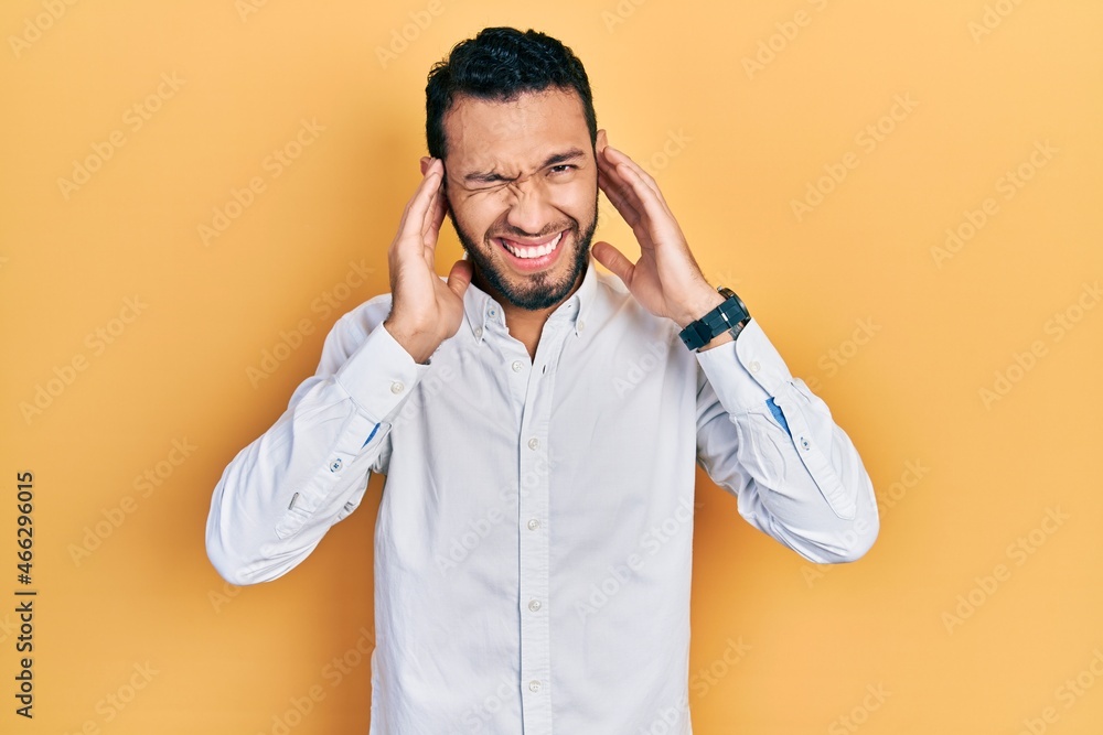 Hispanic man with beard wearing business shirt covering ears with fingers with annoyed expression for the noise of loud music. deaf concept. Hispanic man with beard wearing business shirt covering ears with fingers with annoyed expression for the noise of loud music. deaf concept.