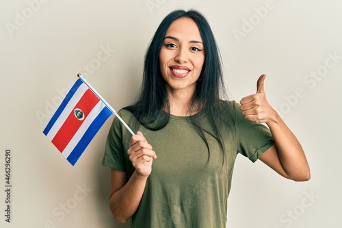 Young hispanic girl holding costa rica flag smiling happy and positive, thumb up doing excellent and approval sign