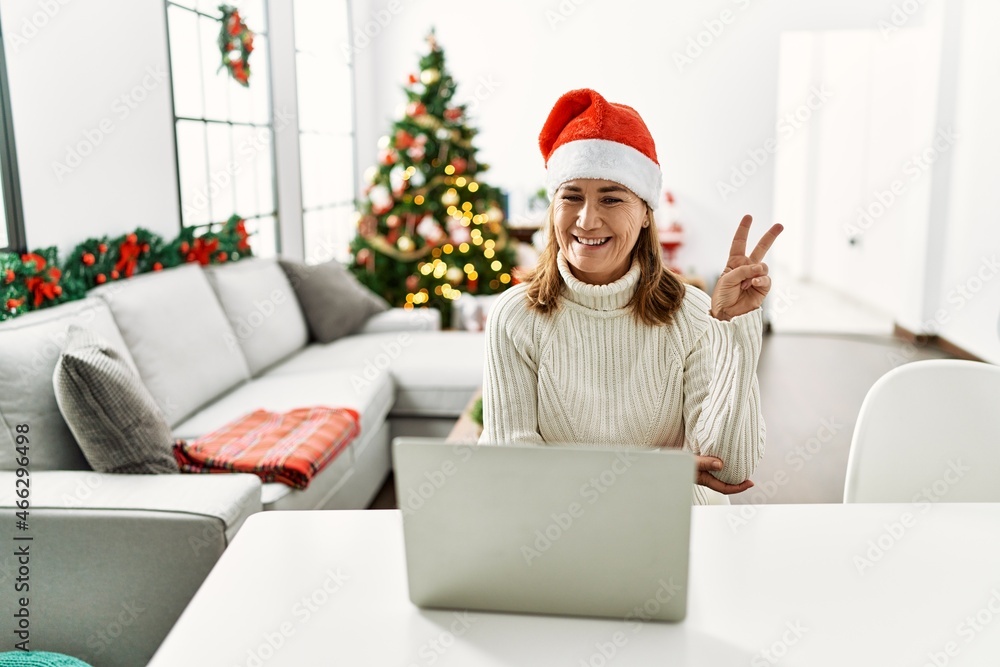 Middle age woman wearing santa claus hat using laptop smiling with happy face winking at the camera doing victory sign. number two. Middle age woman wearing santa claus hat using laptop smiling with happy face winking at the camera doing victory sign. number two.