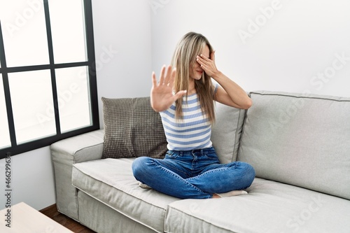 Asian young woman sitting on the sofa at home covering eyes with hands and doing stop gesture with sad and fear expression. embarrassed and negative concept.
