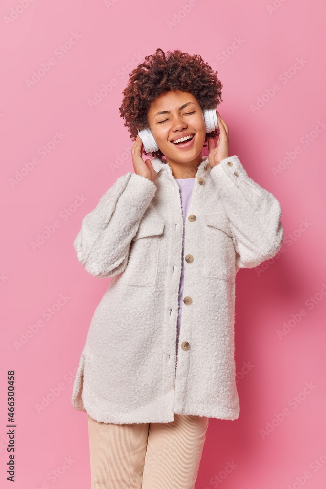 Vertical shot of happy woman meloman keeps eyes closed smiles joyfully listens favorite music in wireless headphones wears fur coat and trousers isolated over pink background. People and hobby Vertical shot of happy woman meloman keeps eyes closed smiles joyfully listens favorite music in wireless headphones wears fur coat and trousers isolated over pink background. People and hobby
