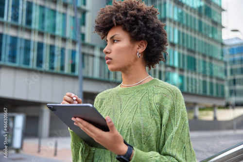 Talented curly female designer with stylus and tablet draws sketches or stickers for social networks while walks outdoors in urban place wears casual green jumper smartwatch has distance job photo