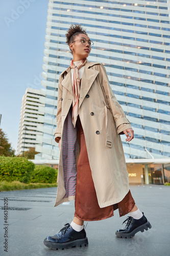 Full body shot of stylish woman dressed in coat and shoes turns back notices something behind walks outdoors against high skyscrapers spends free time in big metropolian city. Urban style concept photo