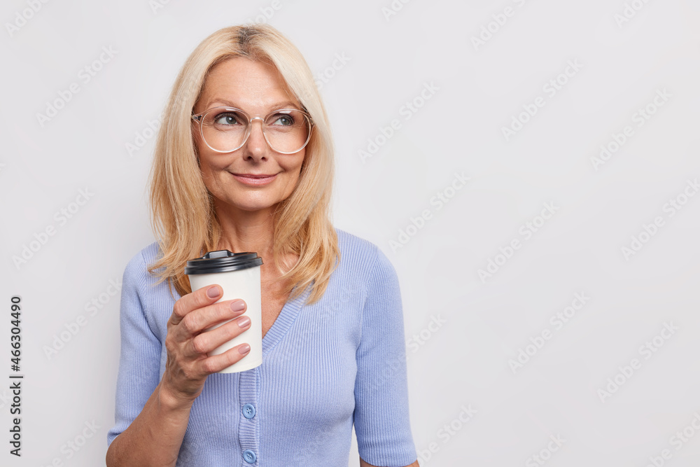 Indoor shot of blonde middle aged European woman with dreamy expression drinks takeaway coffee enjoys aromatic drink wears transparent glasses blue jumper isolated over white background copy space Indoor shot of blonde middle aged European woman with dreamy expression drinks takeaway coffee enjoys aromatic drink wears transparent glasses blue jumper isolated over white background copy space