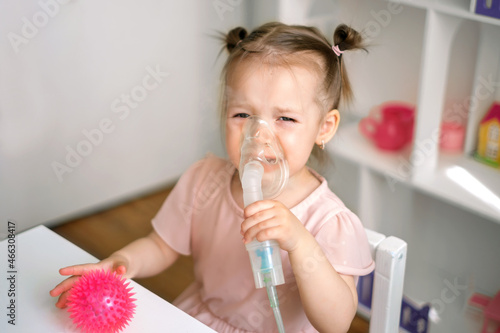 A baby holds a breathing mask and cries while Inhalations. Children's viral diseases. Inhalations for runny nose, respiratory diseases and астме. Children's nebulizer for inhalation.