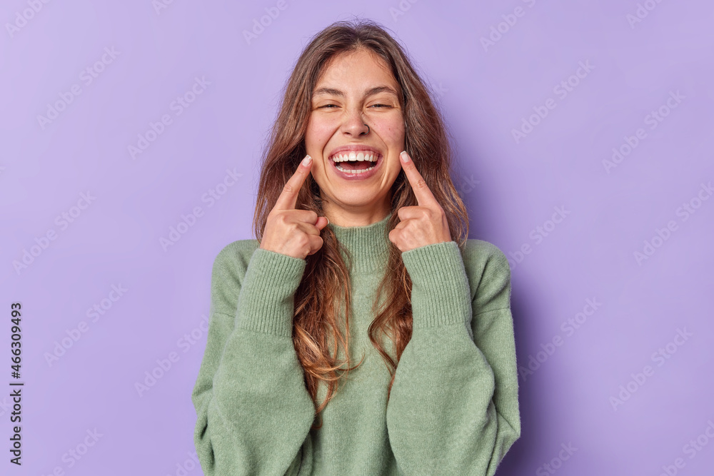 Happy positive dark haired young woman laughs positively points index fingers at toothy smile dressed in casual jumper isolated over purple background. People happy emotions and feelings concept Happy positive dark haired young woman laughs positively points index fingers at toothy smile dressed in casual jumper isolated over purple background. People happy emotions and feelings concept