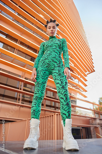 Konstfotografi Below view of fashionable teenage girl dressed in green jumpsuit wears white shoes has trendy hairstyle poses against modern building looks confident at camera
