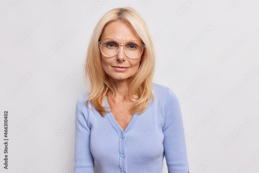 Fototapeta premium Portrait of serious blonde European woman looks directly at camera has minimal makeup wears round spectacles and casual blue jumper isolated over white background has self confident expression
