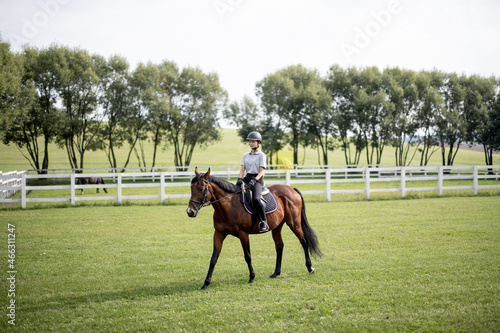 Female horseman riding brown Thoroughbred horse on green meadow in countryside. Concept of rural resting and leisure. Green tourism. Young smiling european woman. Beautiful landscape at sunny day