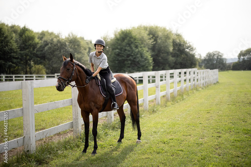 Female horseman riding brown Thoroughbred horse on green meadow in countryside. Concept of rural resting and leisure. Green tourism. Young smiling european woman. Beautiful landscape at sunny day © rh2010