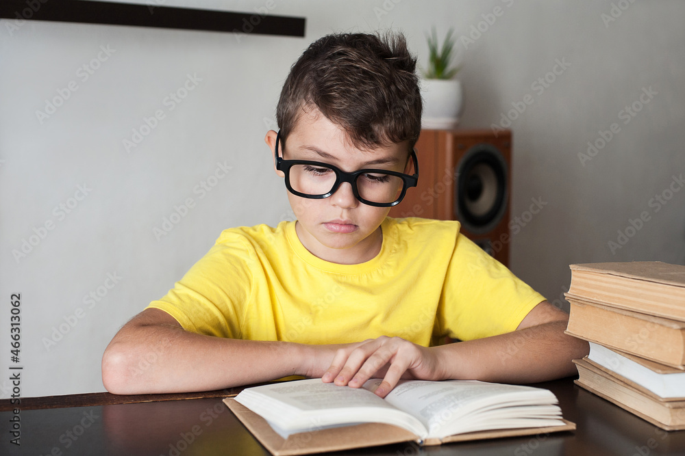a boy with glasses and a yellow T-shirt reads books at the table at home. Home education a boy with glasses and a yellow T-shirt reads books at the table at home. Home education