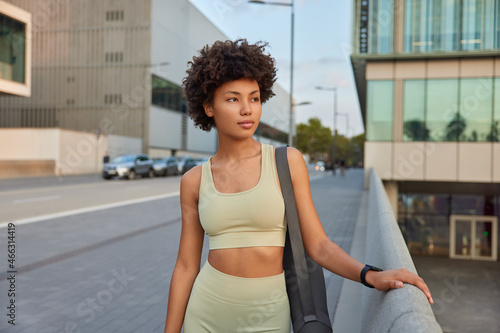Sport recreation and healthy lifestyle concept. Pensive curly haired woman dressed in tracksuit poses on urban road carries karemat going to practice pilates exercises with couch looks away.