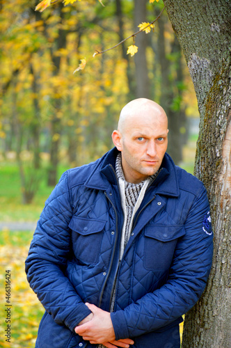 A handsome young man stands near a tree in an autumn park. portrait of a brutal man seriously looking at the camera