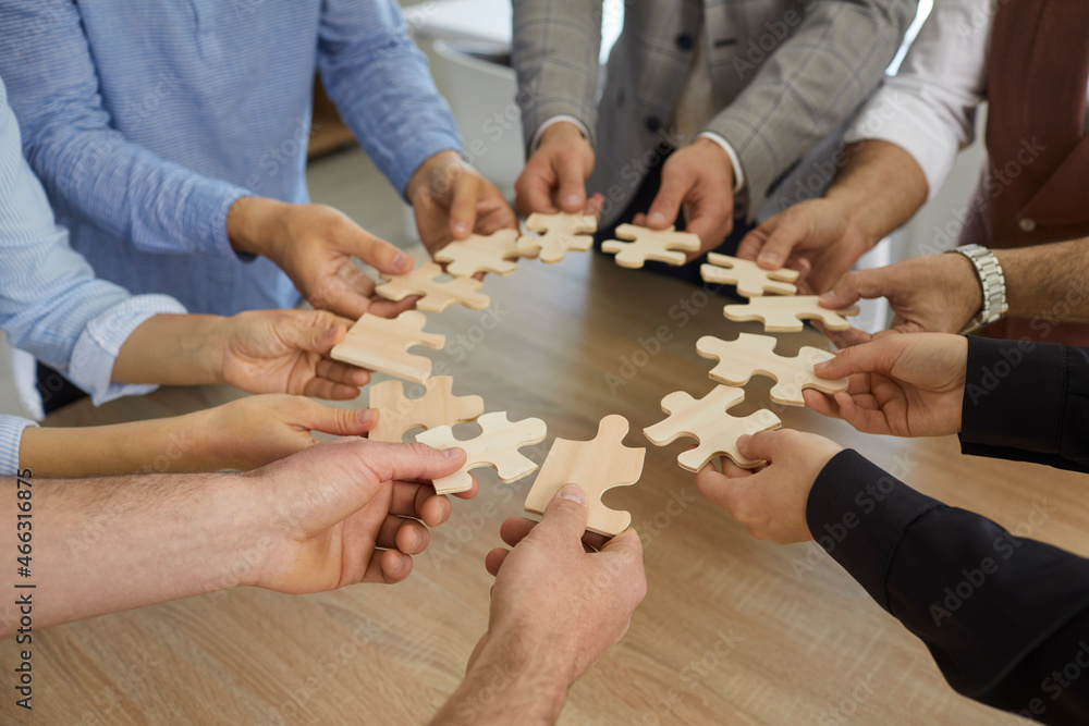 Team of business people standing around office table joining pieces of jigsaw puzzle, close up, closeup shot. Group of entrepreneurs cooperating and forming coalition or alliance. Teamwork concept Team of business people standing around office table joining pieces of jigsaw puzzle, close up, closeup shot. Group of entrepreneurs cooperating and forming coalition or alliance. Teamwork concept