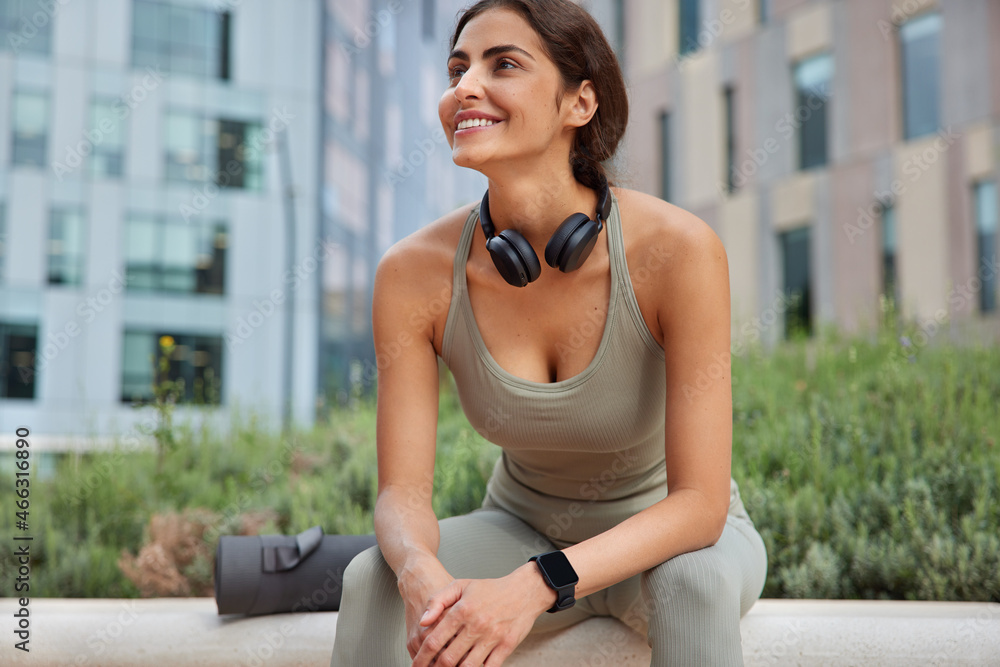 Outdoor shot of satisfied sportswoman rests after cardio training or practicing pilates exercises dressed in sportswear uses headphones smartwatch and karemat poses against urban background.