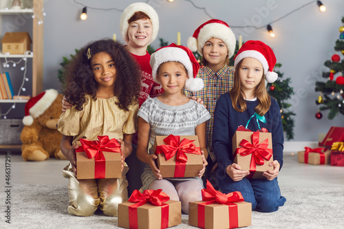 Group of happy smiling multiracial children looking at camera and holding presents tied with red bows, sitting in cozy decorated living-room on Christmas morning at home or during party with friends