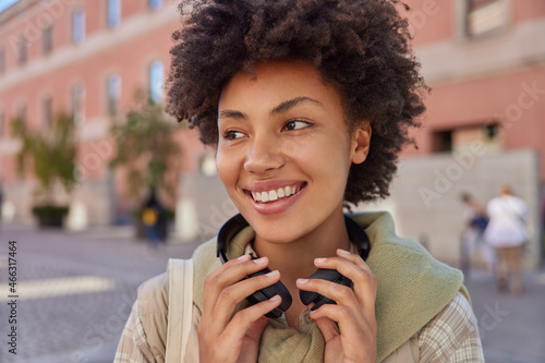 Close up shot of happy curly haired woman wears headphones around neck smiles joyfully strolls outside enjoys spare time poses against blurred background. People hobby and lifestyle concept.