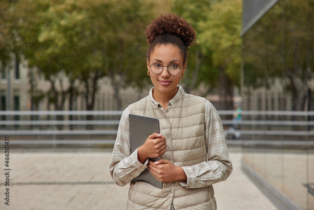 Fototapeta premium Photo of attractive curly haired student dressed in casual clothes holds modern tablet looks directly at camera poses against blurred background in city. People technology and lifestyle concept