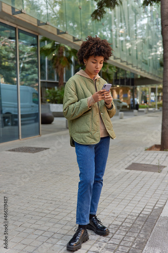 Outdoor shot of serious woman dressed in stylish clothing uses cellular for making quick payment texts in social media chats watches videos online on gadget stands on city pavement near shopping mall