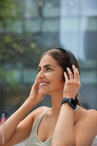 Outdoor shot of happy sporty woman puts on wireless headphones listens music wears werable smartwatch for tracking fitness activity enjoys goos weather for training poses against blurred background