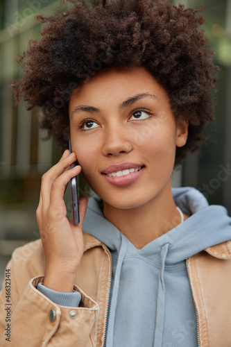 Vertical shot of beautiful curly young woman concentrated somewhere has telephone conversation keeps smartphone near ear wears hoodie and jacket poses against blurred background talks in roaming