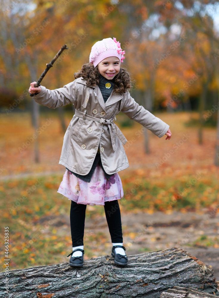 Fototapeta premium Portrait of a girl in an autumn park. The child is standing on the trunk of a tree and playing with a twig
