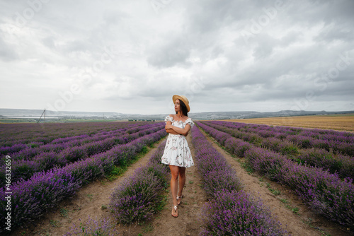 A young beautiful girl in a delicate dress and hat walks through a beautiful field of lavender and enjoys the aroma of flowers. Vacation and beautiful nature.