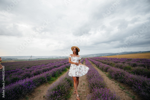 Fototapeta Naklejka Na Ścianę i Meble -  A young beautiful girl in a delicate dress and hat walks through a beautiful field of lavender and enjoys the aroma of flowers. Vacation and beautiful nature.
