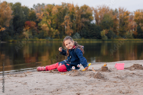 Fototapeta Naklejka Na Ścianę i Meble -  Girl is sitting on sandy beach. Happy little girl in autumn clothes plays on river bank and enjoys warm day in nature. 