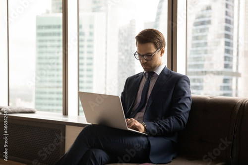 Concentrated young businessman ceo executive manager in formal wear and glasses working on online project on computer, analyzing sales statistics sitting on sofa in modern office with cityscape view.