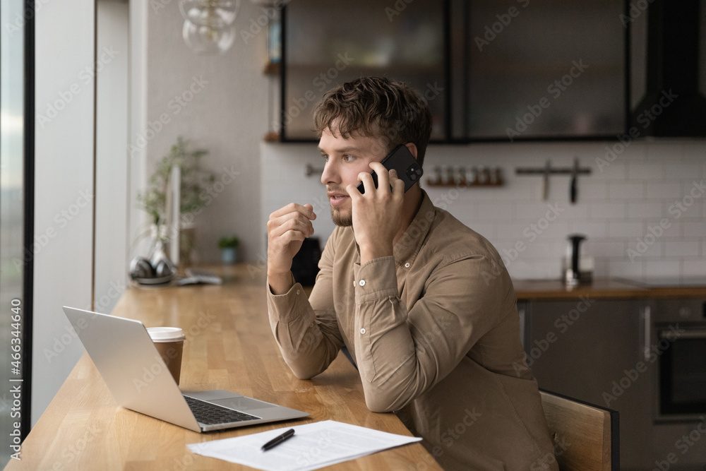 Concentrated young male manager holding phone call conversation, working on laptop sitting at table giving distant professional consultation to client, discussing online project, multitasking concept.