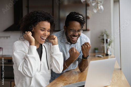 Bild auf Leinwand Overjoyed laughing young african american family couple looking at laptop screen, reading email with amazing news, celebrating online lottery gambling betting auction win, internet success concept