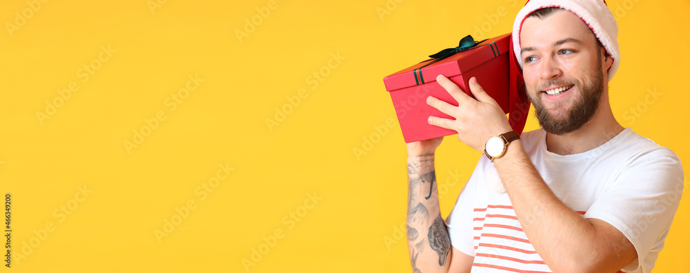 Happy man in Santa hat holding Christmas gift box on yellow background with space for text - obrazy, fototapety, plakaty 