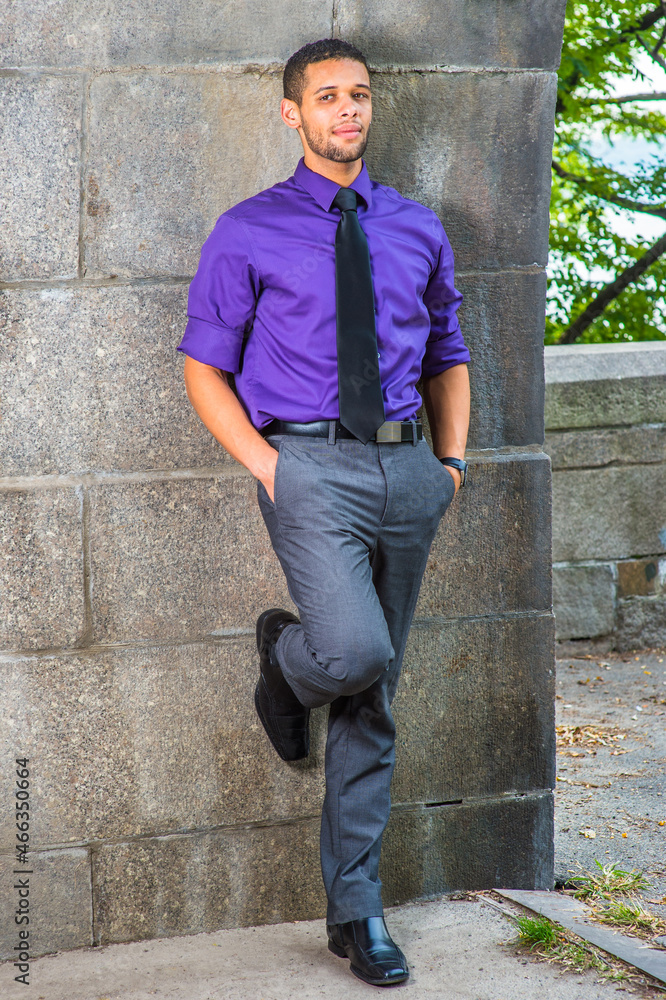 Dressing in a purple shirt, gray pants, a black tie, leather shoes, two hands putting in pockets, one foot touching the wall, a young businessman is leaning against the wall, relaxing.
