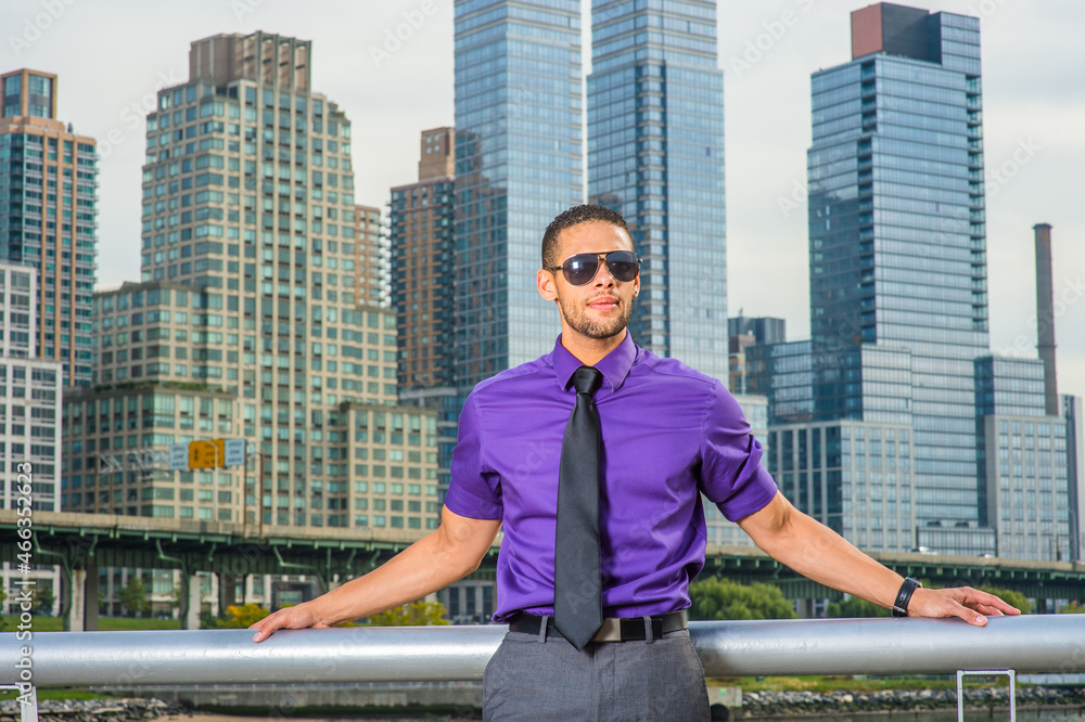 Dressing in a purple shirt, gray pants and a black tie, wearing a sunglasses, a young handsome businessman with a little beard and mustache is standing outside a busy business district..