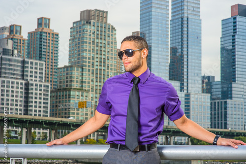 Dressing in a purple shirt, gray pants and a black tie, wearing a sunglasses, a young handsome businessman with a little beard and mustache is standing outside a busy business district...