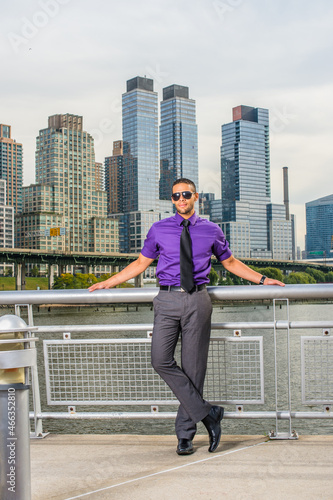 Dressing in a purple shirt, gray pants, a black tie, leather shoes, wearing a sunglasses, a young businessman with a little beard and mustache is standing outside a busy business district, relaxing..