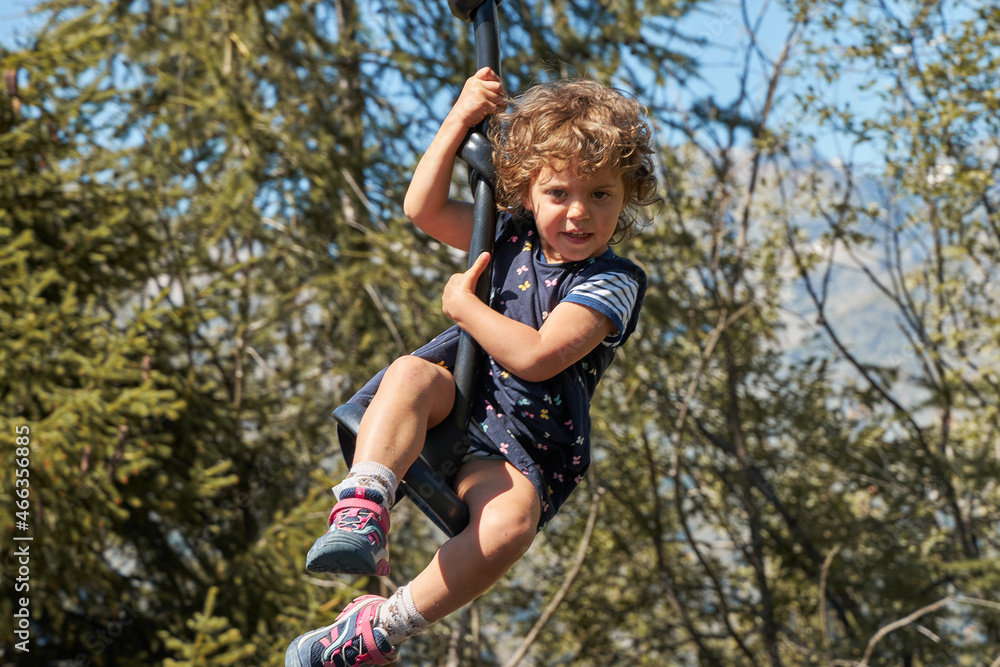 A happy curly-haired girl rides on a high swing in the mountains. Light curly hair. He looks at the camera. A happy curly-haired girl rides on a high swing in the mountains. Light curly hair. He looks at the camera.