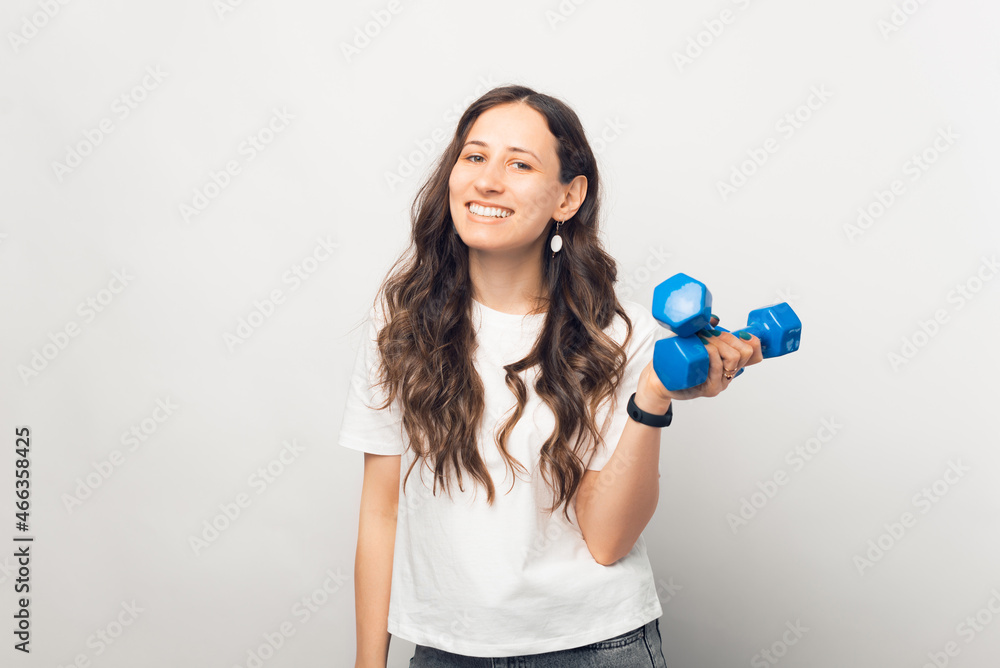 Young smiling brunette is holding some blue dumbbells. She is wearing white T-shirt. Young smiling brunette is holding some blue dumbbells. She is wearing white T-shirt.