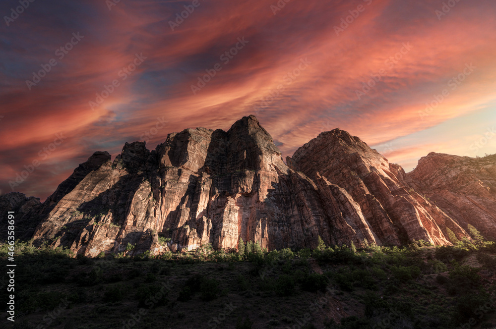 Fototapeta premium The setting sunlight shines upon a mountain range of jagged rocks in Zion National Park. The sky is colorful with red, pink and orange clouds.