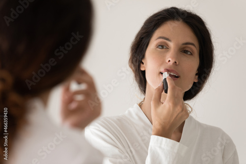 Female in bathrobe enjoy self-grooming in front of mirror holds lipstick applying color on lips looks at reflection feels satisfied, use decorative cosmetics for women beauty, morning routine concept