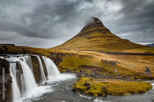 Closeup landscape photo of Kirkjufell mountain, Iceland, with three-tiered waterfall in foreground. Cloudy gray sky, brown and yellow vegetation.