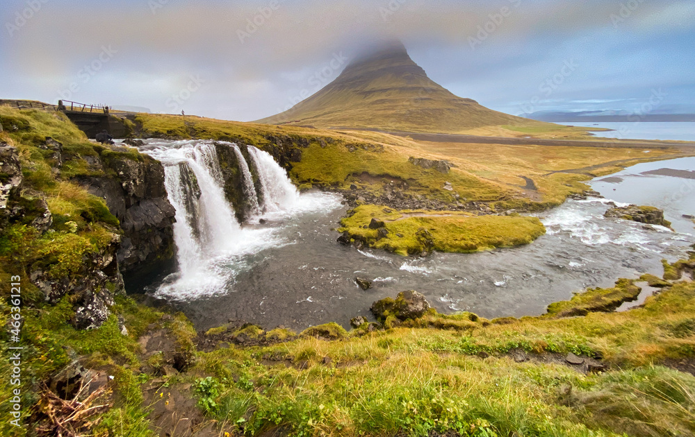 Kirkjufell mountain in Iceland. Three-tiered waterfall in foreground, mountain in background, top if mountain in clouds.