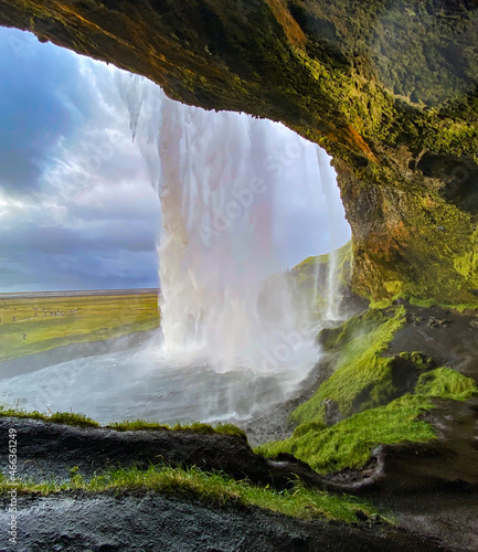 Rear cave view of Seljalandsfoss waterfall in Iceland. Cave rim has highlights or orange and green.  Patches of light on waterfall from late afternoon sunlight.