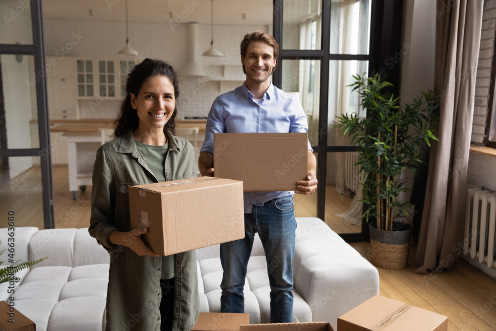Beautiful 35s spouses pose in modern living room with cardboard boxes in hands, young family relocating to new own first house bringing personal belongings feel happy. Bank mortgage, tenancy concept Beautiful 35s spouses pose in modern living room with cardboard boxes in hands, young family relocating to new own first house bringing personal belongings feel happy. Bank mortgage, tenancy concept