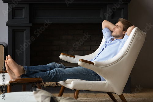 Serene young man put hands behind head, feet on footstool, relaxing in leather comfortable armchair in luxury home with fireplace. No stress, fatigue relieve, furniture store ad, peace of mind concept photo