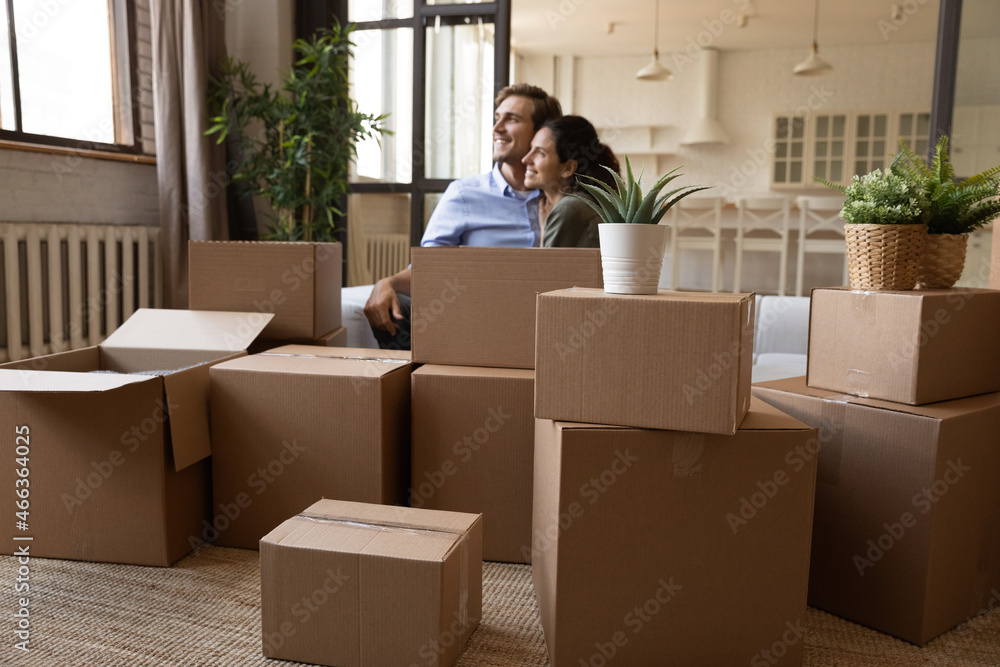 Young serene couple relaxing on sofa near heap of cardboard boxes with personal belongings on relocation day, take break, feel happy on moving day to new first own house. Bank loan, mortgage concept Young serene couple relaxing on sofa near heap of cardboard boxes with personal belongings on relocation day, take break, feel happy on moving day to new first own house. Bank loan, mortgage concept